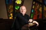 Fr. Dave Pivonka sitting in a church in front of stain glass windows.