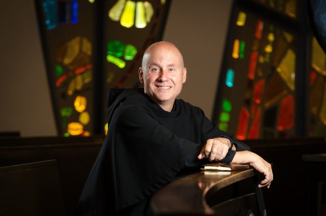 Fr. Dave Pivonka sitting in a church in front of stain glass windows.