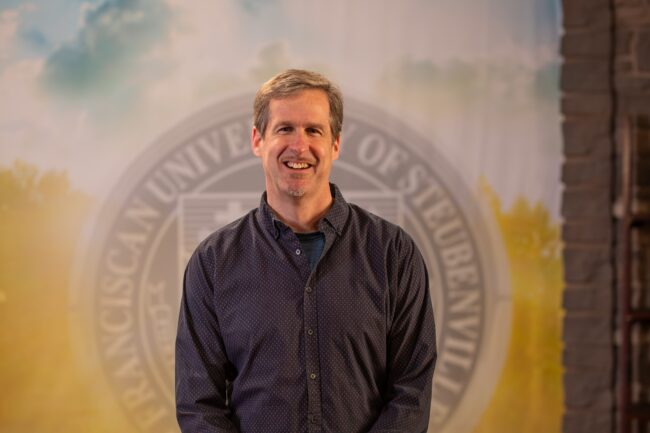 To Web – Dr. Daniel Kuebler FEB2025–2 Dr. Dan Kuebler standing in front of the Franciscan University of Steubenville seal.