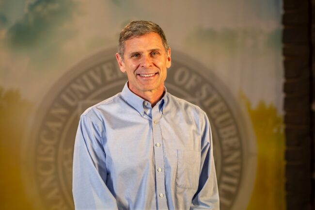 To Web – Dr. Jeffrey Rohde FEB2025 –7 Dr. Jeffrey Rohde standing in front of university seal.