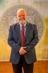 Dr. Petroc Willey standing in front of a university seal.