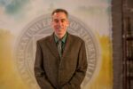 Dr. Ron Bolster standing in front of the Franciscan University of Steubenville seal.
