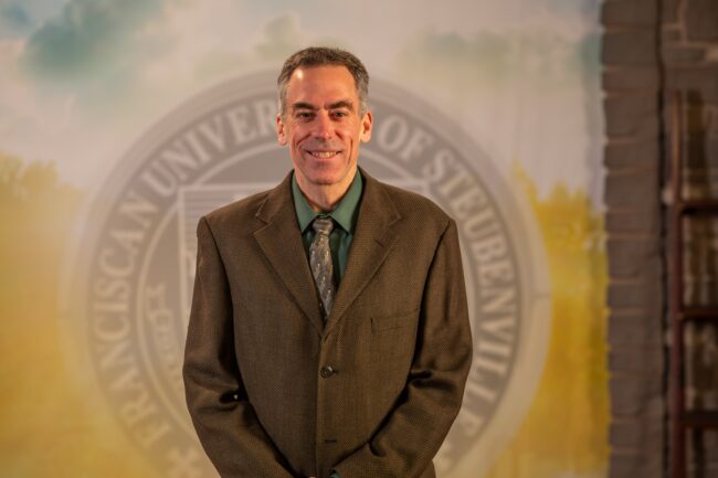 To Web – Dr. Ron Bolster FEB2025 –8 Dr. Ron Bolster standing in front of the Franciscan University of Steubenville seal.