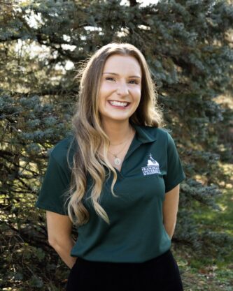 Megan Case headshot Portrait of Megan Case wearing a Franciscan University of Steubenville polo shirt, standing outdoors in front of trees.