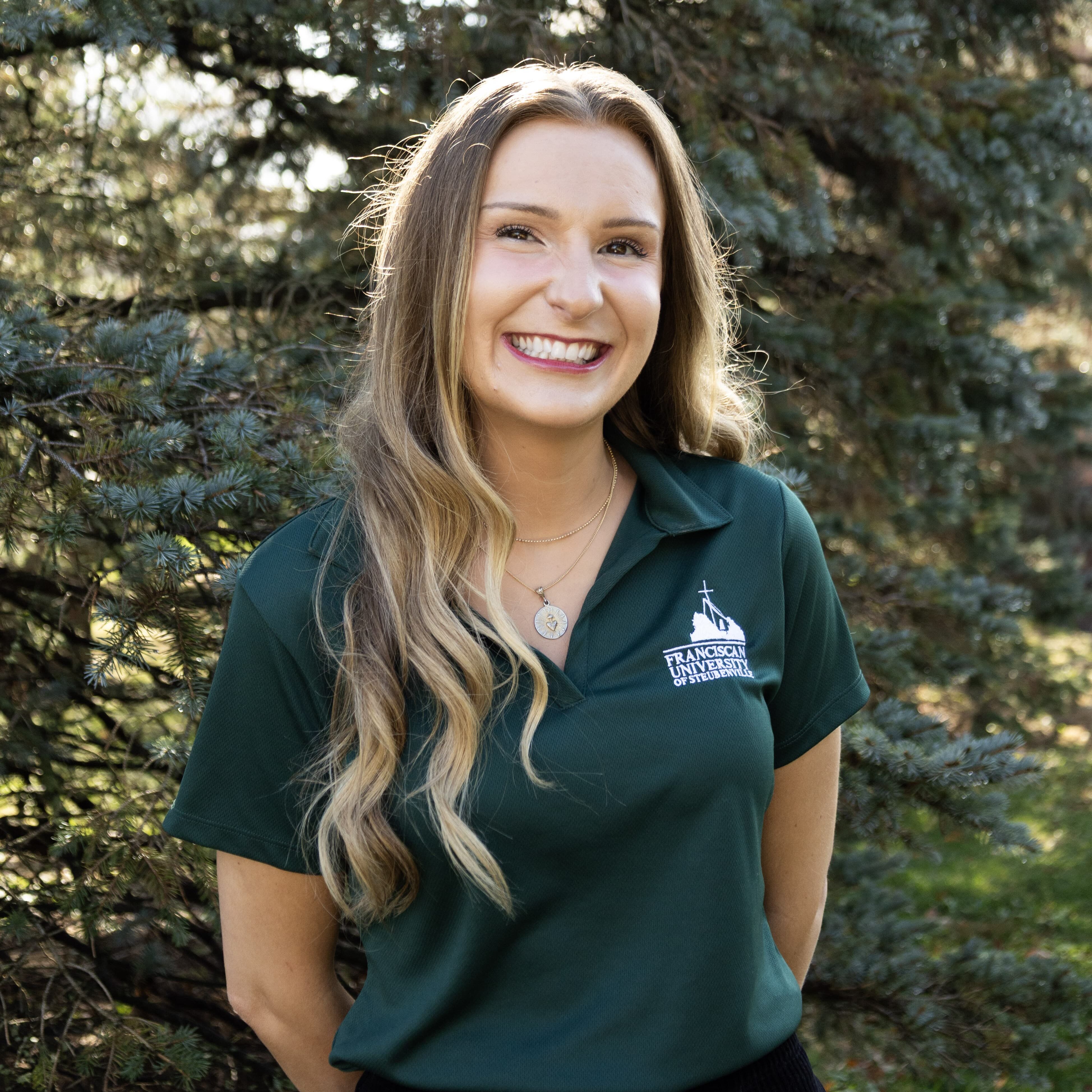 Megan Case headshot Portrait of Megan Case wearing a Franciscan University of Steubenville polo shirt, standing outdoors in front of trees.