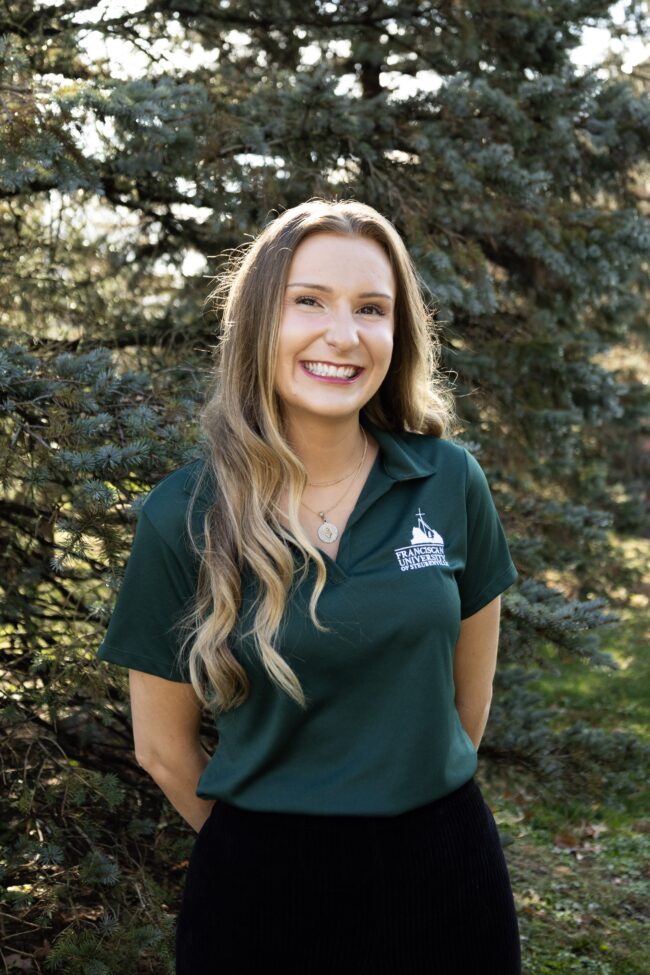 Portrait of Megan Case wearing a Franciscan University of Steubenville polo shirt, standing outdoors in front of trees.