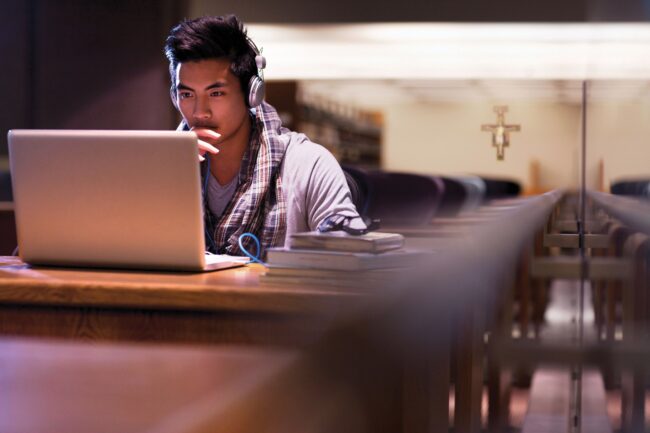 A student wearing headphones studies on a laptop at a wooden table in a library, with a cross visible in the background.