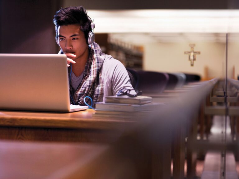 A student wearing headphones studies on a laptop at a wooden table in a library, with a cross visible in the background.
