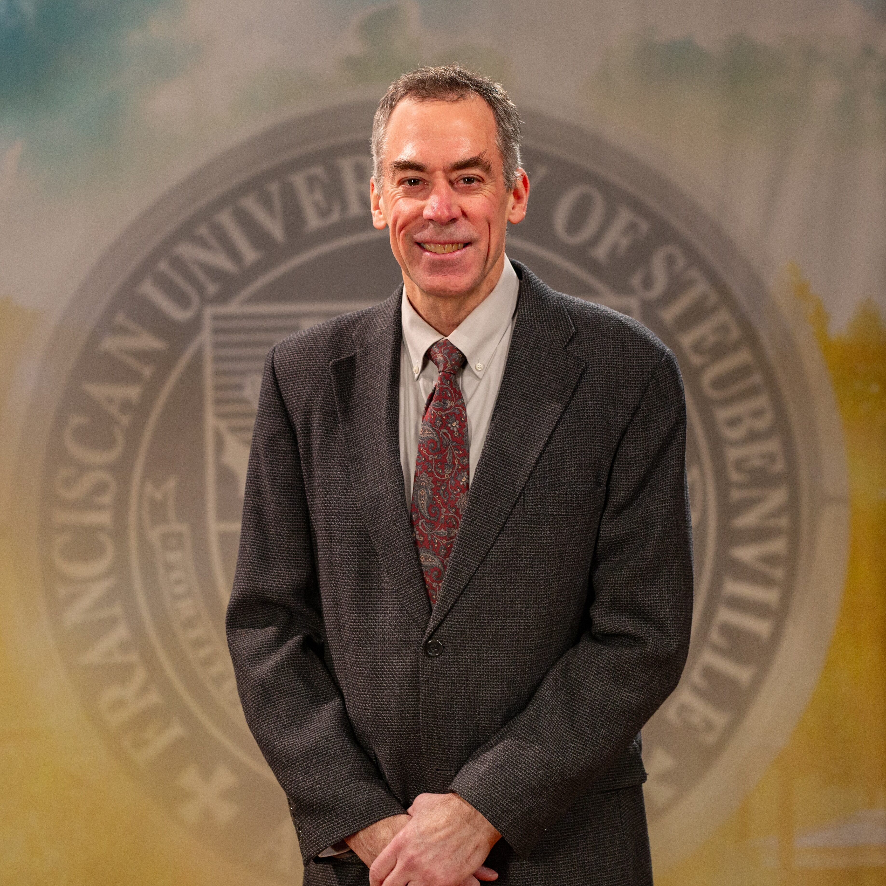 Dr. Ron Bolster_Faculty Portrait_Spring2026_-2816 Portrait of Dr. Ron Bolster standing in front of the Franciscan University of Steubenville seal.