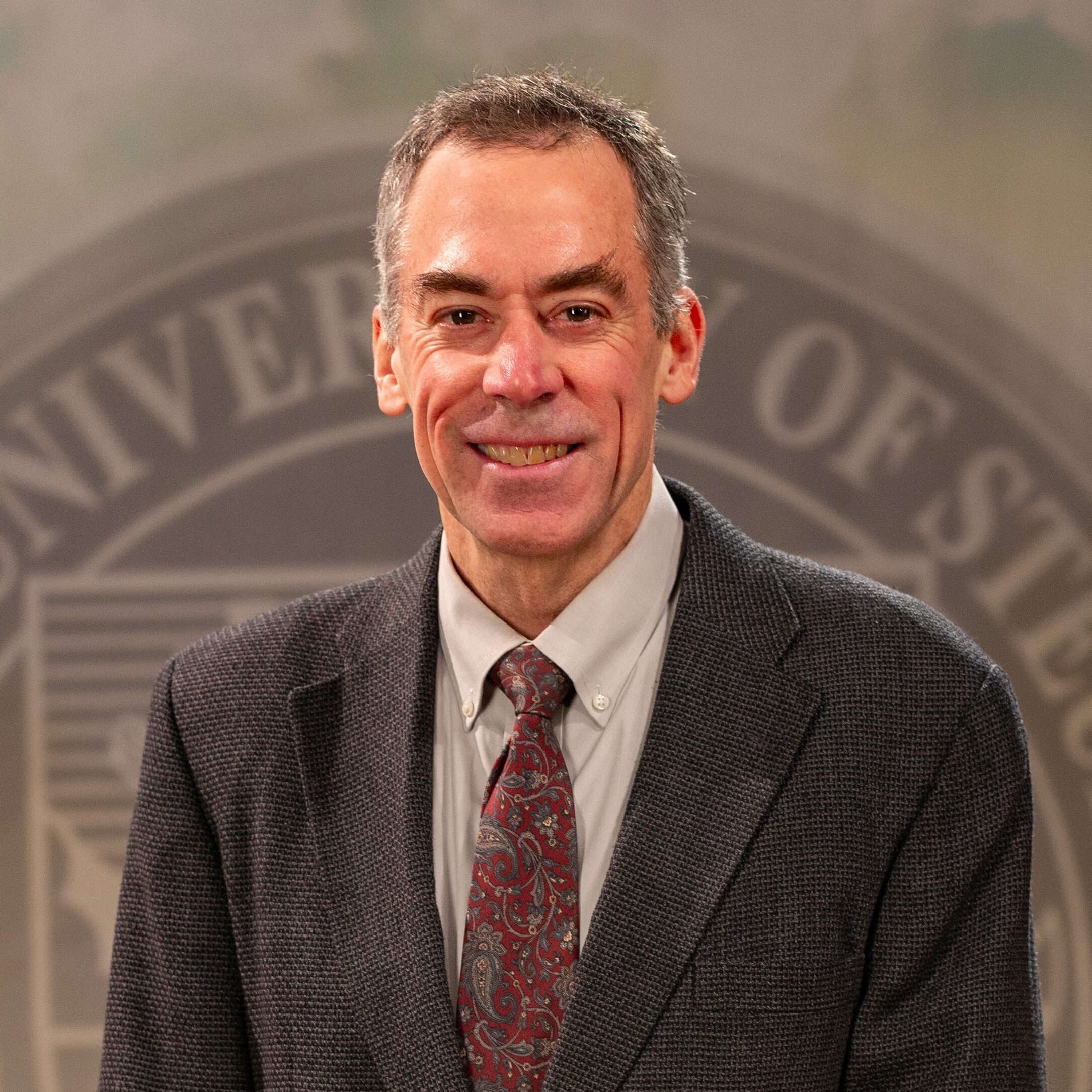 Dr. Ron Bolster_Faculty Portrait_Spring2026_-2816 Portrait of Dr. Ron Bolster standing in front of the Franciscan University of Steubenville seal.