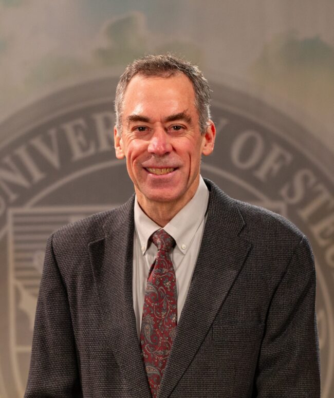 Portrait of Dr. Ron Bolster standing in front of the Franciscan University of Steubenville seal.