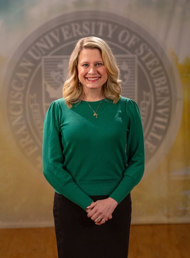 Faculty portrait of Dr. Rebecca Rook standing in front of the Franciscan University of Steubenville seal.